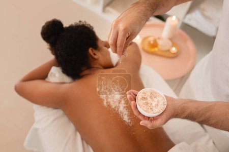 L'image montre un gros plan des mains saupoudrant un gommage exfoliant sur une femme afro-américaine cliente lors d'un traitement spa