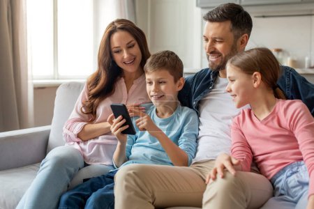 Foto de Sonriente familia sentada en el sofá en el acogedor interior de la casa como un niño les muestra algo en un teléfono inteligente - Imagen libre de derechos
