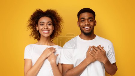 Kind hearted young african-american woman and man keep both palms on chest, express gratitude and good feeling, orange background