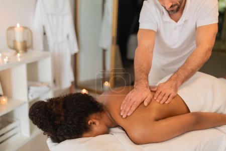 Photo for A masseur gives a relaxing back massage to African American woman lying on a spa bed, environment looks serene - Royalty Free Image