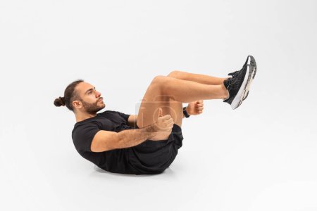 Energetic man in black athletic clothing performing a floating crunch exercise against a plain white backdrop