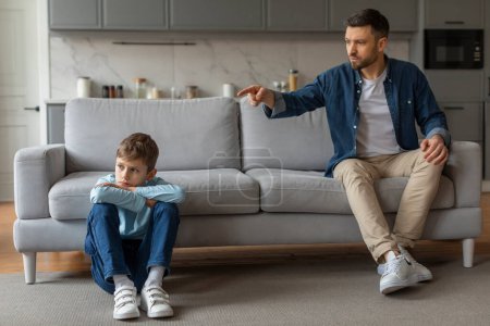 Hombre con atuendo casual padre parece estar regañando a un joven hijo enfurruñado sentado en el suelo junto a un sofá en un hogar moderno