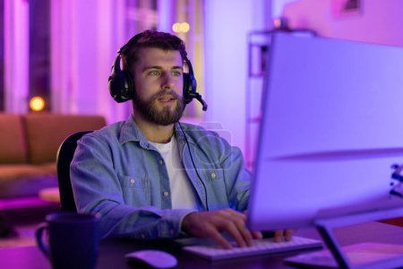 In a room with neon lights, a focused man uses a headset while using a computer with a serious expression