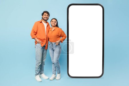 Téléchargez les photos : Homme et femme indiens souriants posant à côté d'un énorme smartphone, indiquant la connectivité ou des applications, espace de copie de maquette - en image libre de droit
