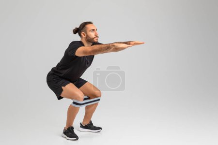 Ein fokussierter Mann macht Kniebeugen mit einem Widerstandsband um die Knie in einem neutralen Rahmen auf grauem Hintergrund, Kopierraum