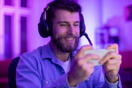 Photo for A smiling bearded guy gamer wearing a headset enjoys using his smartphone in a neon-lit gaming setup - Royalty Free Image