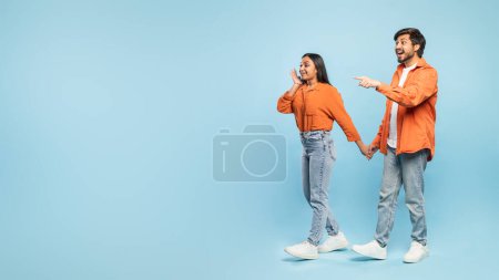 Photo for Indian man and woman in matching orange shirts pointing to the side in surprise on a light blue background - Royalty Free Image
