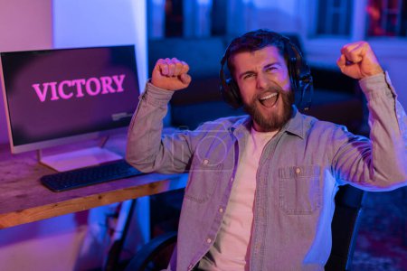 Gamer hombre mostrando la expresión extática con la VICTORIA en la pantalla, las manos levantadas, y la atmósfera de luz azul