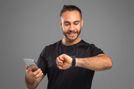 A guy in casual clothing checks his smartwatch while holding a smartphone, tracking his workout, grey background