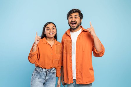 Photo for Indian man and woman both in orange, excitedly pointing up with bright expressions on a blue background - Royalty Free Image