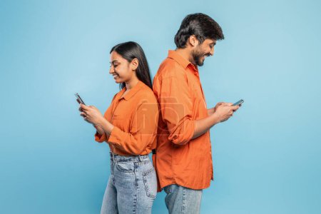 Foto de Un joven indio y una mujer se paran espalda con espalda, absortos en sus móviles sobre un fondo azul, destacando la desconexión a pesar de la proximidad - Imagen libre de derechos