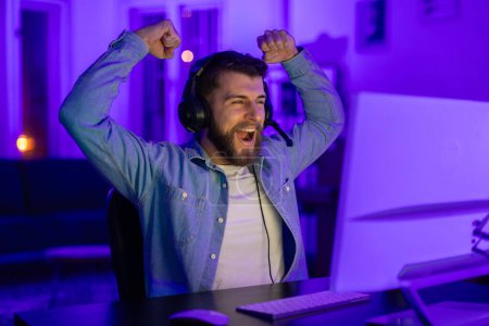 Gamer guy celebrates a victory with enthusiastic cheers, surrounded by blue light in a gaming setup