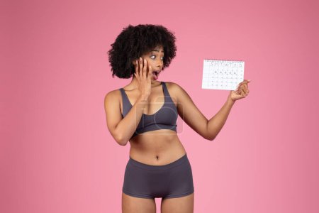 Foto de Mujer afroamericana estresada en un sujetador deportivo mirando el calendario del ciclo menstrual y tocándose la cara en un telón de fondo rosa - Imagen libre de derechos