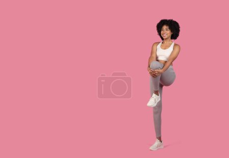 Fit young african american woman performing a leg stretch against a pink backdrop, illustrating flexibility and routine