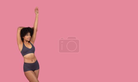 Téléchargez les photos : Une jeune femme afro-américaine en forme prend une pose ludique dans un ensemble sportif de deux pièces, sur un fond rose vif - en image libre de droit