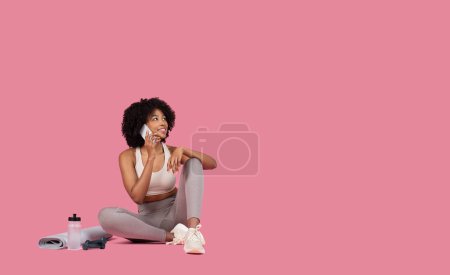Photo for A young African American woman in sportswear sits with her phone, looking thoughtful, against a pink background - Royalty Free Image
