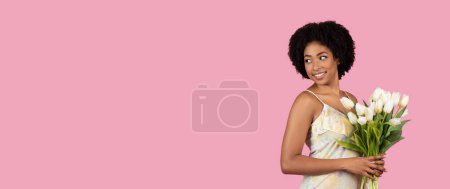Foto de Mujer afroamericana en un vestido de verano sosteniendo tulipanes mira hacia otro lado, sobre un fondo rosa liso, espacio para copiar - Imagen libre de derechos