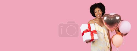 Photo for A playful African American woman carrying a white gift box and colorful heart-shaped balloons on a pink background - Royalty Free Image
