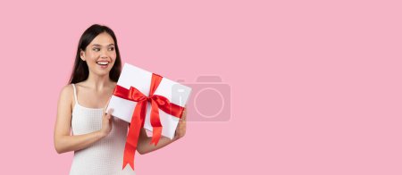 Smiling woman holds a large white gift box with a red ribbon, space for text on the left side on a pink backdrop