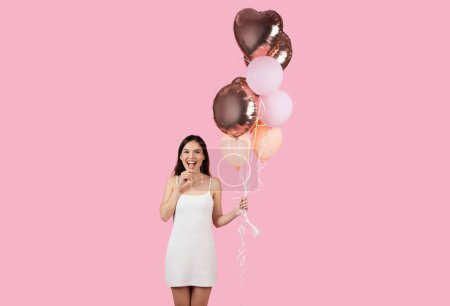 Photo for An excited young woman in white dress holds balloons, expression of fun and celebration on a pink background - Royalty Free Image