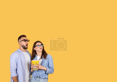 Photo for Two friends man and woman in 3D glasses express shock, sharing a popcorn snack on a yellow backdrop - Royalty Free Image
