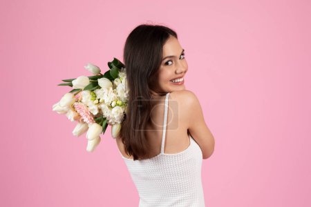 A smiling young woman in a white tank top looks over her shoulder holding a bouquet, on a pink backdrop
