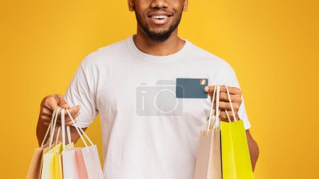 Foto de Alegre hombre afroamericano milenario con coloridas bolsas de papel y tarjeta de crédito, fondo de estudio naranja - Imagen libre de derechos