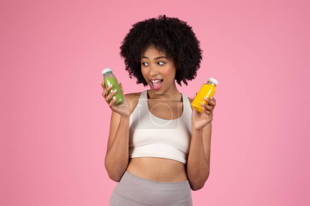 Foto de Mujer afroamericana feliz comparando dos botellas de jugos nutritivos coloridos - Imagen libre de derechos