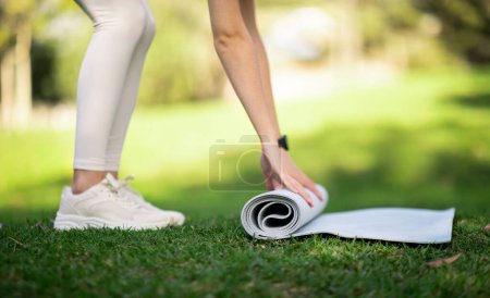 Photo for Close-up of a person athlete in white leggings and sneakers rolling up a yoga mat in a lush green park, indicating preparation or completion of a workout, outside, cropped - Royalty Free Image