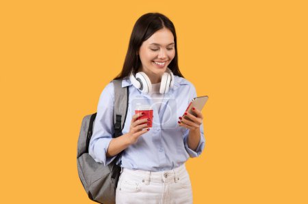 Photo for Young woman student texting on phone and holding a red coffee cup, with backpack on yellow background - Royalty Free Image