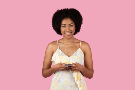 Photo for Smiling young African American woman using a cell phone, wearing a summer dress against a pink background - Royalty Free Image