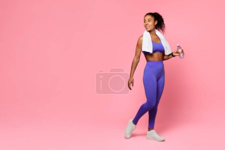 Foto de Mujer afroamericana en traje de gimnasio azul sonríe mientras camina, sosteniendo una botella de agua y una toalla, sobre un fondo rosa - Imagen libre de derechos