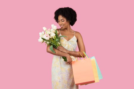 Téléchargez les photos : Belle dame afro-américaine sentant un bouquet de fleurs, avec des sacs à provisions sur un fond rose - en image libre de droit