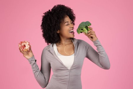 A smiling african american woman is torn between a choice of a healthy broccoli and a tempting doughnut
