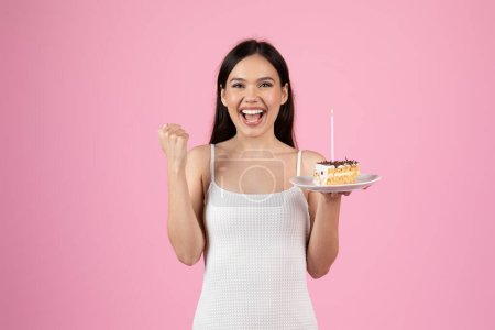 Young woman shows excitement with a cake in one hand and a victory gesture in another on pink background