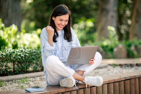 Photo for A delighted young woman shows a gesture of success after good news on her laptop, studying at public park - Royalty Free Image