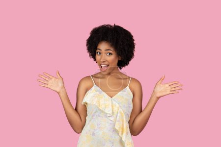 Happy pretty African American woman in dress with open arms and a surprised expression on pink background