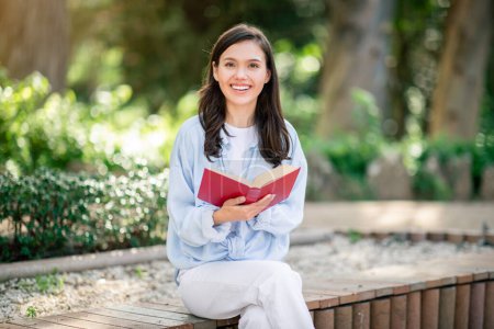 Photo for Enthusiastic smiling european young reader sits on a park bench with a beaming smile, engrossed in a captivating red book, enjoying the peaceful outdoor environment, outside - Royalty Free Image