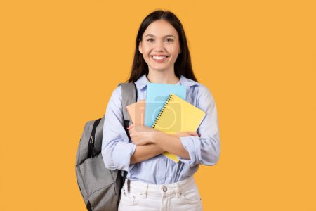 Téléchargez les photos : Jeune femme adulte étreignant des cahiers colorés, souriant, avec un sac à dos, posant sur fond de studio jaune - en image libre de droit