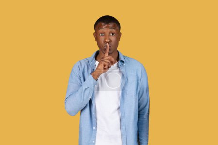 African American young man places a finger over his lips signaling for silence or secrecy against a yellow backdrop