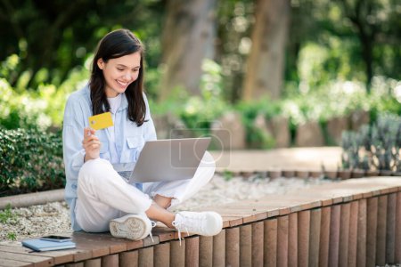 Foto de Joven europea sonriente sentada al aire libre con una computadora portátil, sosteniendo una tarjeta de crédito, probablemente haciendo una compra en línea en un ambiente relajado y soleado del parque, afuera. Venta, banca - Imagen libre de derechos