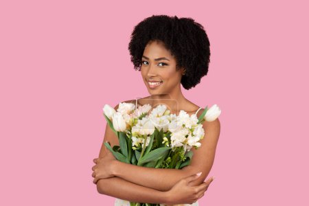 Content African American woman hugging a bunch of white flowers, looking to the side on pink background