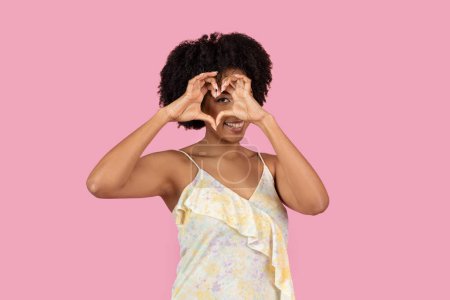 A confident young African American woman stands with heart shaped gesture, against a vivid pink background, wearing a yellow summer dress