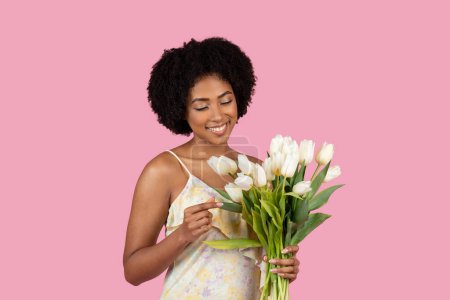 Foto de Una joven y alegre mujer afroamericana con el pelo rizado sosteniendo un ramo fresco de tulipanes blancos sobre un telón de fondo rosa - Imagen libre de derechos
