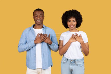 Foto de El hombre y la mujer afroamericanos están agradecidos poniendo las manos en sus pechos, expresando agradecimiento o amor sobre un fondo amarillo - Imagen libre de derechos