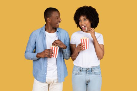 Téléchargez les photos : Couple afro-américain engagé et joyeux appréciant le pop-corn et bavardant sur le jaune, allant au cinéma - en image libre de droit