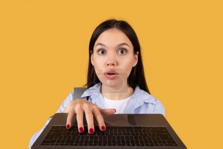 Foto de Mujer joven con expresión sorprendida mirando sobre un portátil abierto, sobre un fondo amarillo, escribiendo en el teclado de la computadora - Imagen libre de derechos