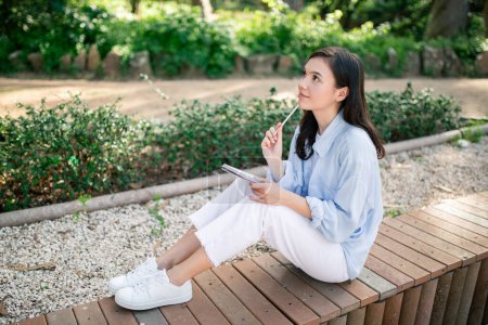 Téléchargez les photos : Une jeune femme européenne réfléchie avec un crayon sur les lèvres s'assoit sur une plateforme en bois dans un parc, réfléchissant à ses notes, immergée dans la réflexion au milieu du calme de la nature, à l'extérieur - en image libre de droit