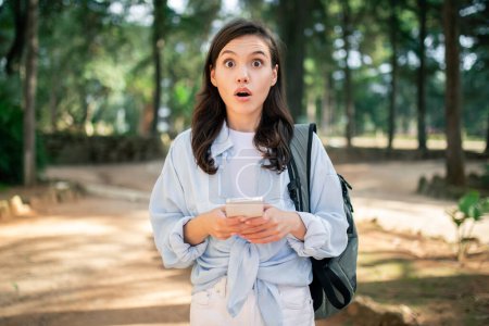 A shocked young woman student reacts to a surprising message on her phone outdoors at public park