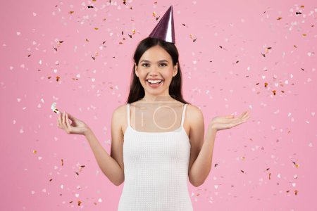 Photo for Smiling woman wears a party hat with colorful confetti falling, festive party atmosphere on pink background - Royalty Free Image
